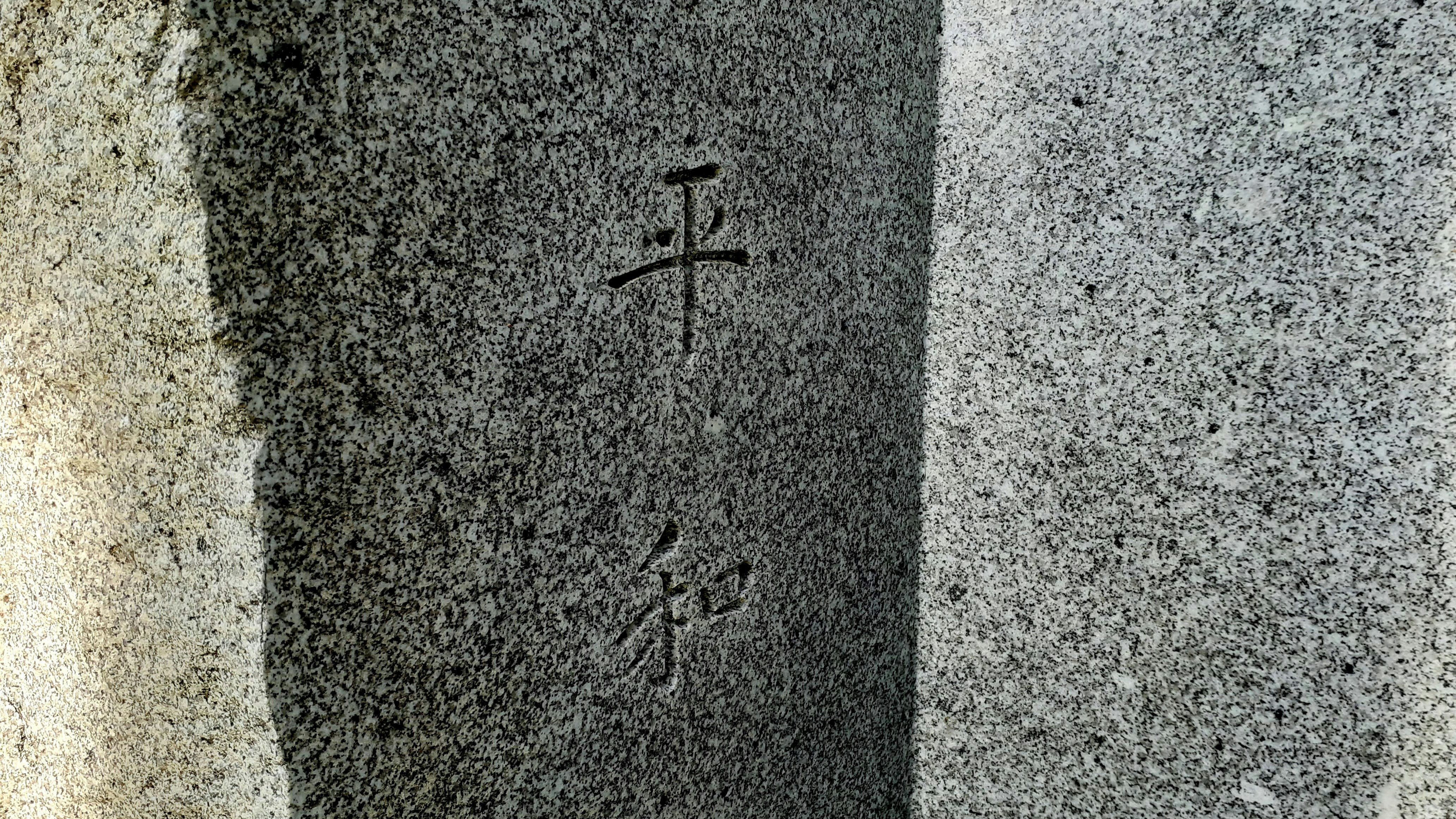 A close-up view of a granite stone surface with engraved Chinese or Japanese characters. The stone appears textured and has variations in light and shadow, giving it a three-dimensional feel.