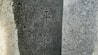 A close-up view of a granite stone surface with engraved Chinese or Japanese characters. The stone appears textured and has variations in light and shadow, giving it a three-dimensional feel.
