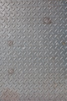A textured metal surface featuring a repetitive diamond plate pattern, typically used for industrial flooring. The metallic texture is highlighted by the shiny, reflective surface of the material.