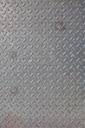 A textured metal surface featuring a repetitive diamond plate pattern, typically used for industrial flooring. The metallic texture is highlighted by the shiny, reflective surface of the material.