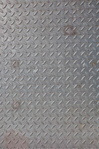 A textured metal surface featuring a repetitive diamond plate pattern, typically used for industrial flooring. The metallic texture is highlighted by the shiny, reflective surface of the material.
