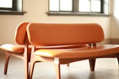 Two wooden chairs with orange leather upholstery placed closely together in a well-lit room with sunlight streaming in through the windows. The chairs have curved backs and seats, and the overall design appears modern and minimalistic.