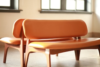 Two wooden chairs with orange leather upholstery placed closely together in a well-lit room with sunlight streaming in through the windows. The chairs have curved backs and seats, and the overall design appears modern and minimalistic.