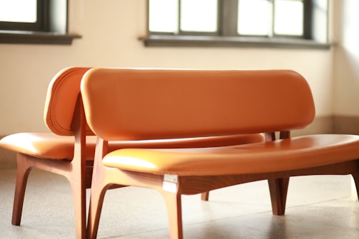 Two wooden chairs with orange leather upholstery placed closely together in a well-lit room with sunlight streaming in through the windows. The chairs have curved backs and seats, and the overall design appears modern and minimalistic.