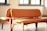 Two wooden chairs with orange leather upholstery placed closely together in a well-lit room with sunlight streaming in through the windows. The chairs have curved backs and seats, and the overall design appears modern and minimalistic.