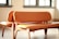 Two wooden chairs with orange leather upholstery placed closely together in a well-lit room with sunlight streaming in through the windows. The chairs have curved backs and seats, and the overall design appears modern and minimalistic.