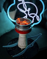 A hookah with glowing coals sits on a dark surface. In the background, there is a neon light shaped like a stylized animal, possibly a rabbit or a hare, with a blue outline. The setup creates a moody and atmospheric scene with contrasting colors.