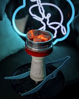 A hookah with glowing coals sits on a dark surface. In the background, there is a neon light shaped like a stylized animal, possibly a rabbit or a hare, with a blue outline. The setup creates a moody and atmospheric scene with contrasting colors.
