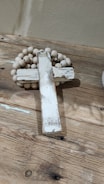 A rustic, weathered wooden cross is placed on a wooden surface, accompanied by a string of wooden beads. The cross has a distressed white paint finish, giving it a vintage look. The beads are light-colored, complementing the overall earthy tones of the scene.