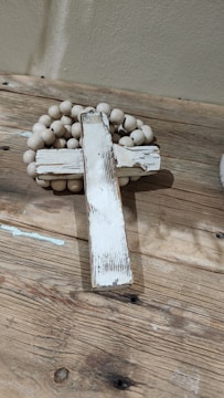 A rustic, weathered wooden cross is placed on a wooden surface, accompanied by a string of wooden beads. The cross has a distressed white paint finish, giving it a vintage look. The beads are light-colored, complementing the overall earthy tones of the scene.