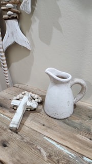 A rustic wooden table holding a weathered white ceramic pitcher and a decorative cross made of distressed wood, accompanied by a beaded garland. Above the table hangs a decorative piece resembling a fish tail made from wood and stones.