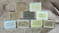 Nine rectangular blocks of soap are neatly arranged on a textured burlap fabric background. Each soap is embossed with the words 'SAVONS ARTHUR.' The soaps vary in shades, including earthy tones and pastel greens.