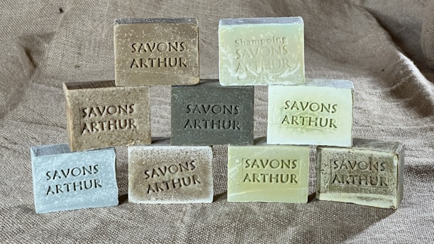 Nine rectangular blocks of soap are neatly arranged on a textured burlap fabric background. Each soap is embossed with the words 'SAVONS ARTHUR.' The soaps vary in shades, including earthy tones and pastel greens.