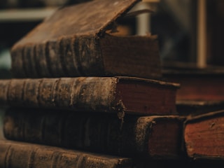 A stack of old, worn books with dark, textured covers and faded red page edges, conveying a sense of antiquity and history.
