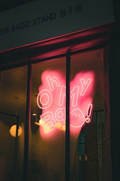 A neon sign reading 'Oh My Bao!' is displayed on a storefront window. The sign glows pink, casting a warm reflection on the glass. Inside, modern pendant lights are visible, and the setting suggests a trendy and inviting atmosphere.