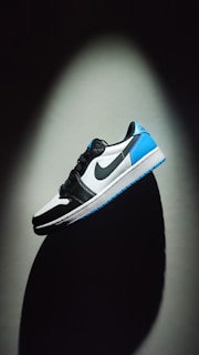 A sneaker is prominently displayed against a dark background. The shoe features a color scheme of black, white, and bright blue. The Nike logo is visible on the side. The lighting creates a dramatic shadow beneath the shoe, enhancing its contours and design.