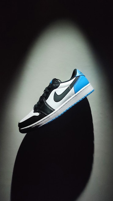 A sneaker is prominently displayed against a dark background. The shoe features a color scheme of black, white, and bright blue. The Nike logo is visible on the side. The lighting creates a dramatic shadow beneath the shoe, enhancing its contours and design.