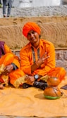 A person is sitting on a mat outdoors, wearing a vibrant orange outfit and turban, holding up a peace sign. Another person partially visible is beside them, also in orange attire. A traditional musical instrument lies on the mat.