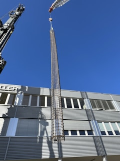 A large crane is lifting a cylindrical metallic structure in front of a modern industrial building under a clear blue sky. The building has a facade with horizontal lines and numerous windows. Shadows from the crane and structure are cast on the building's surface.