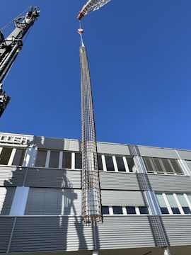 A large crane is lifting a cylindrical metallic structure in front of a modern industrial building under a clear blue sky. The building has a facade with horizontal lines and numerous windows. Shadows from the crane and structure are cast on the building's surface.