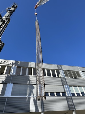 A large crane is lifting a cylindrical metallic structure in front of a modern industrial building under a clear blue sky. The building has a facade with horizontal lines and numerous windows. Shadows from the crane and structure are cast on the building's surface.