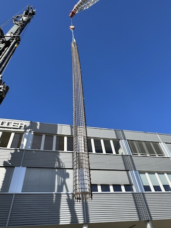 A large crane is lifting a cylindrical metallic structure in front of a modern industrial building under a clear blue sky. The building has a facade with horizontal lines and numerous windows. Shadows from the crane and structure are cast on the building's surface.