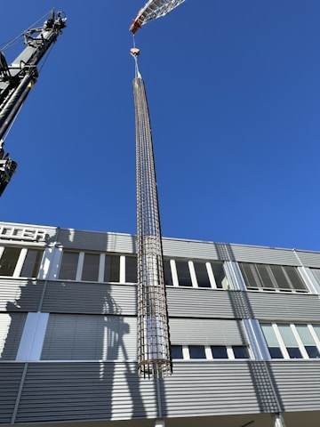 A large crane is lifting a cylindrical metallic structure in front of a modern industrial building under a clear blue sky. The building has a facade with horizontal lines and numerous windows. Shadows from the crane and structure are cast on the building's surface.