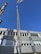 A large crane is lifting a cylindrical metallic structure in front of a modern industrial building under a clear blue sky. The building has a facade with horizontal lines and numerous windows. Shadows from the crane and structure are cast on the building's surface.