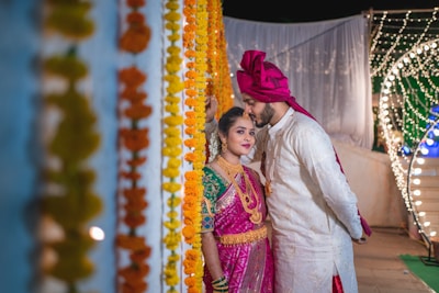A couple dressed in traditional Indian attire partakes in a wedding ceremony. The woman is wearing a vibrant pink saree with intricate gold jewelry and vivid green blouse, while the man is dressed in an elegant white sherwani and a pink turban. They stand surrounded by garlands of marigold flowers and strings of lights, creating a festive and intimate atmosphere.