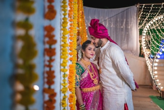 A couple dressed in traditional Indian attire partakes in a wedding ceremony. The woman is wearing a vibrant pink saree with intricate gold jewelry and vivid green blouse, while the man is dressed in an elegant white sherwani and a pink turban. They stand surrounded by garlands of marigold flowers and strings of lights, creating a festive and intimate atmosphere.