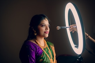 A woman is seated in profile, looking calmly forward as a makeup brush is held near her face. She is adorned in traditional attire with a green and purple saree, gold jewelry, and hair ornaments. A circular ring light illuminates her from the front, creating a warm, focused atmosphere.