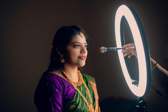 A woman is seated in profile, looking calmly forward as a makeup brush is held near her face. She is adorned in traditional attire with a green and purple saree, gold jewelry, and hair ornaments. A circular ring light illuminates her from the front, creating a warm, focused atmosphere.