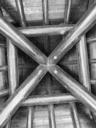 A geometric wooden structure with a cross pattern at its center and beams extending outward. The construction hints at a roof or ceiling framework, showcasing intersecting lines and a symmetrical design.