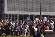 A diverse crowd of people gathered outdoors holding signs and banners advocating for equality and against hate. Several individuals are displaying rainbow flags, indicating support for LGBTQ+ rights. The setting appears to be in an urban area in front of a building with a unique patterned facade.
