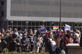 A diverse crowd of people gathered outdoors holding signs and banners advocating for equality and against hate. Several individuals are displaying rainbow flags, indicating support for LGBTQ+ rights. The setting appears to be in an urban area in front of a building with a unique patterned facade.