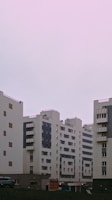Several modern apartment buildings with a minimalist architectural design stand under a pastel pink sky. The buildings feature a combination of white and dark panels with numerous windows and small balconies.