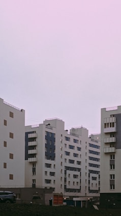 Several modern apartment buildings with a minimalist architectural design stand under a pastel pink sky. The buildings feature a combination of white and dark panels with numerous windows and small balconies.