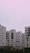 Several modern apartment buildings with a minimalist architectural design stand under a pastel pink sky. The buildings feature a combination of white and dark panels with numerous windows and small balconies.