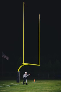 A referee wearing a black and white striped uniform stands on a football field near a tall, yellow goalpost. An American flag is visible in the background, and the sky is dark, suggesting a night game. The referee appears to be signaling with a thumbs-up gesture.