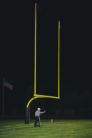A referee wearing a black and white striped uniform stands on a football field near a tall, yellow goalpost. An American flag is visible in the background, and the sky is dark, suggesting a night game. The referee appears to be signaling with a thumbs-up gesture.