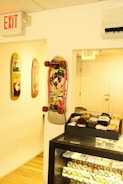 A skateboard with a colorful graphic design is mounted on a wall along with two other skateboards. Below is a glass display case filled with various skateboard wheels and neatly arranged stacks of socks on top. The room has a light wooden floor and is well-lit with bright overhead lighting. Two exit signs are visible, one above a doorway and the other on the ceiling.