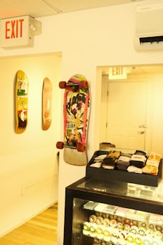 A skateboard with a colorful graphic design is mounted on a wall along with two other skateboards. Below is a glass display case filled with various skateboard wheels and neatly arranged stacks of socks on top. The room has a light wooden floor and is well-lit with bright overhead lighting. Two exit signs are visible, one above a doorway and the other on the ceiling.