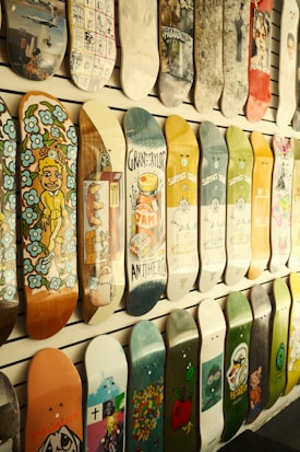 Several skateboard decks are displayed on a wall, each featuring unique and colorful designs. The designs range from abstract patterns, cartoon characters to pop art and advertisements. The decks are arranged in an orderly fashion, creating a visually appealing and artistic display.