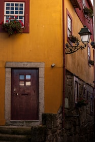 A charming yellow building with a wooden door featuring a number and the letters 'AL'. Red-framed windows are adorned with vibrant flower boxes. A vintage-style lamp post adds an old-world charm to the setting. The stone steps and textured walls suggest a historic or quaint setting.