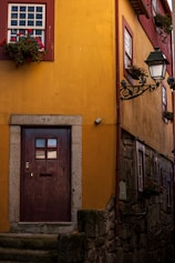A charming yellow building with a wooden door featuring a number and the letters 'AL'. Red-framed windows are adorned with vibrant flower boxes. A vintage-style lamp post adds an old-world charm to the setting. The stone steps and textured walls suggest a historic or quaint setting.