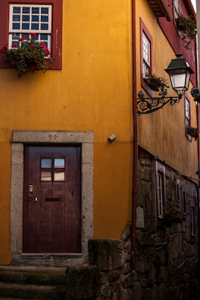 A charming yellow building with a wooden door featuring a number and the letters 'AL'. Red-framed windows are adorned with vibrant flower boxes. A vintage-style lamp post adds an old-world charm to the setting. The stone steps and textured walls suggest a historic or quaint setting.
