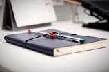 A desk with a closed book in a dark blue cover, laying flat with two pens placed on top, one pen is red and the other green and black. The background includes a blurred calendar and parts of a computer keyboard, lending a tidy and organized atmosphere.