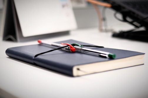 A desk with a closed book in a dark blue cover, laying flat with two pens placed on top, one pen is red and the other green and black. The background includes a blurred calendar and parts of a computer keyboard, lending a tidy and organized atmosphere.