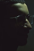 A dark, moody profile of a person with delicate rimless glasses. The lighting is low, casting deep shadows that partially obscure facial features, providing a mysterious and introspective atmosphere.