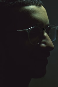 A dark, moody profile of a person with delicate rimless glasses. The lighting is low, casting deep shadows that partially obscure facial features, providing a mysterious and introspective atmosphere.